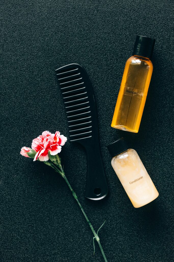 Flat lay of hair care essentials with a comb, bottles, and pink flower on dark background.